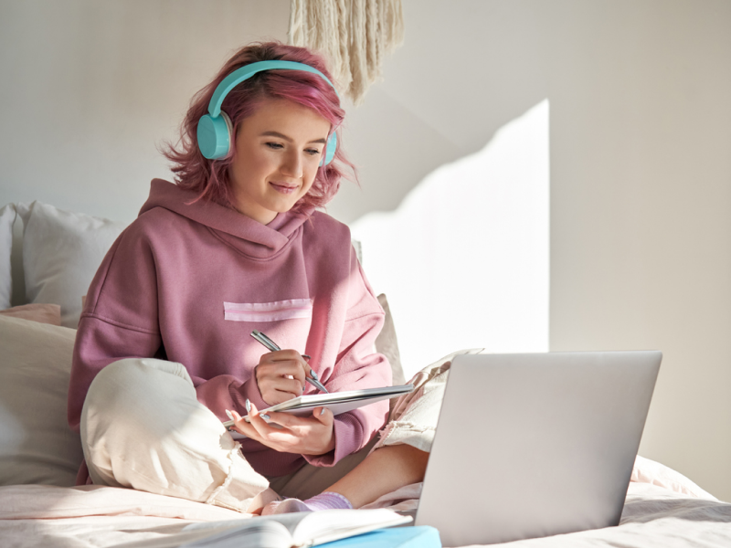 Teen female sitting on her bed in a pink sweatshirt and green headphones. She is in a DBT skills group.