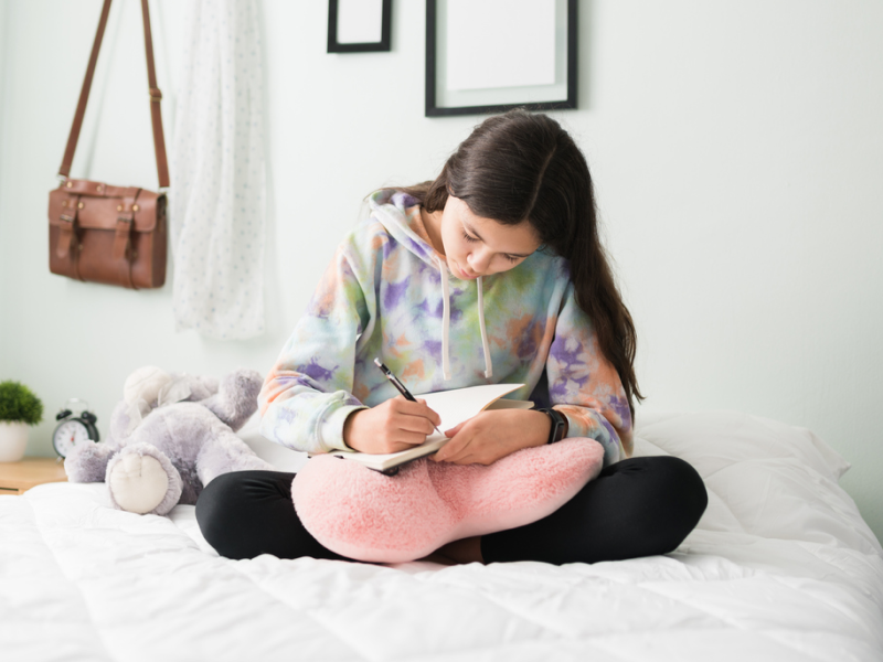 A young person in a tie dye sweatshirt sits on her bed journaling for mental health.