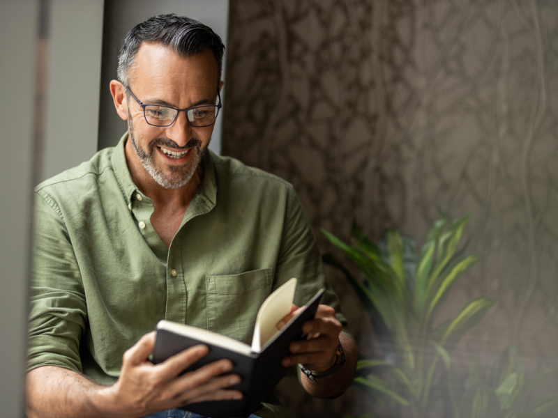 Male therapist in green shirt leaning on a wall reading a book.
