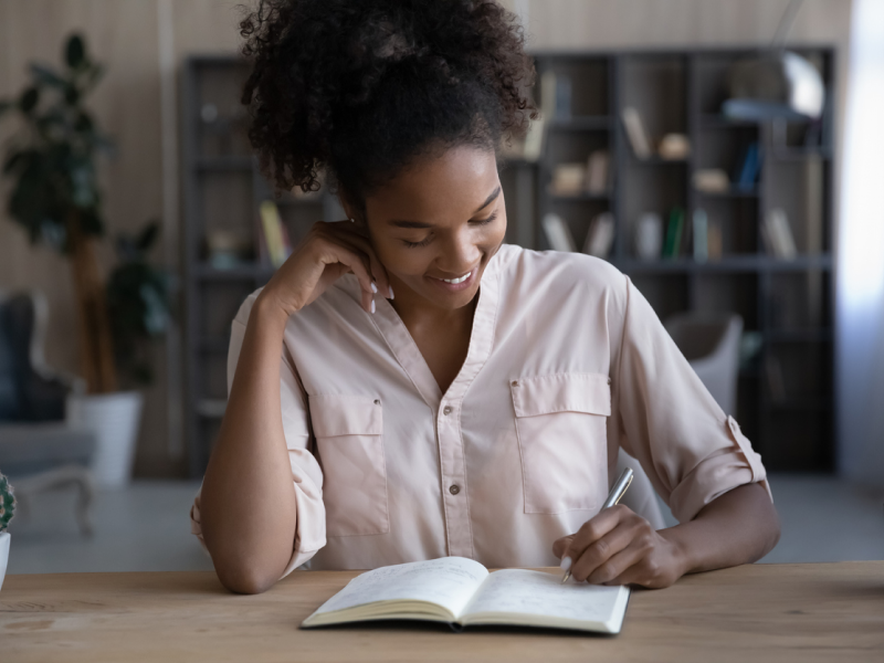 Teen in a pink shirt writing in her journal on the table. Teen is setting mental health goals for herself.