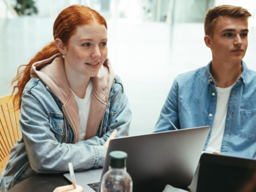 A young woman with red hair sits in front of her computer next to a young man. She is smiling because she is in control of her emotions after using the check the facts DBT skill.