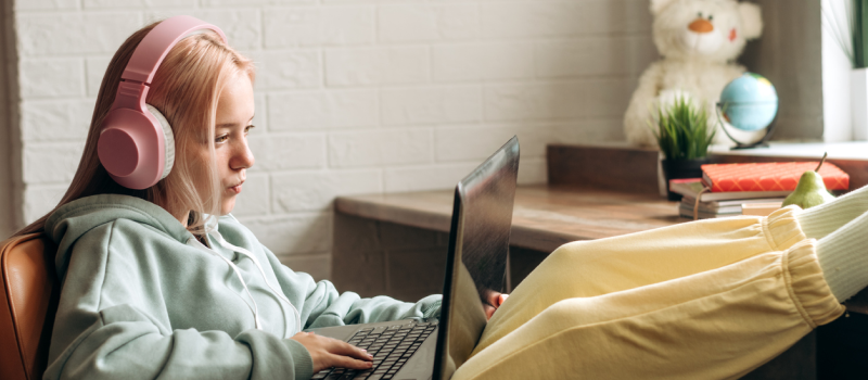 A young woman in pink headphones and a blue sweatshirt sits in a chair at her desk in front of her laptop during a group therapy session where she is learning about the check the facts DBT skill.