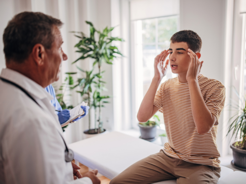 A person in a tan shirt sits in a doctor's office explaining his mental health symptoms as the doctor tries to determine if he is dealing with factitious disorder or malingering.