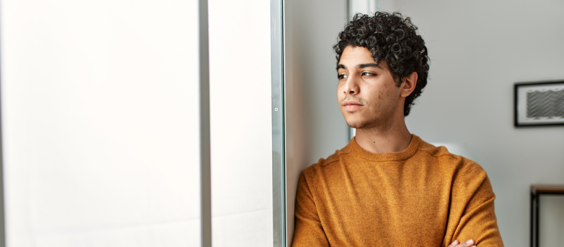 Male young adult in an orange shirt leaning on the wall looking out of the window. The individual was recently diagnosed with anxiety.