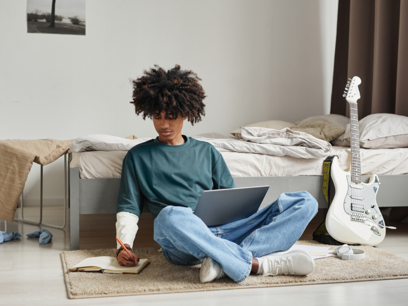 A young person sits on the floor next to his guitar journaling, utilizing the tools he knows to manage his overstimulation.