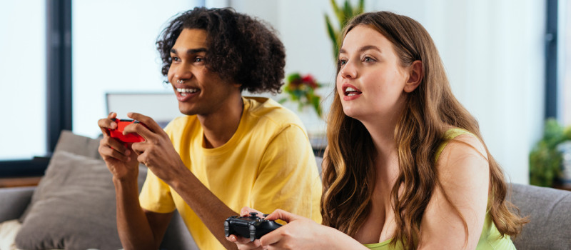 Two young people sit next to each other on a couch playing video games. Gaming is positively impacting their mental health and they are not dealing with internet gaming addiction.
