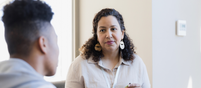 A therapist in a beige shirt and earrings listens to a patient who is considering which form of therapy is best for him.