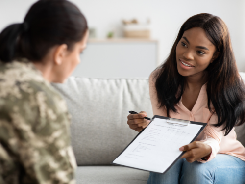 Woman in pink shirt sitting on a couch across from a veteran in a military uniform. The woman in the pink shirt is a counselor helping the veteran understand her depression and anxiety symptoms and seek help.