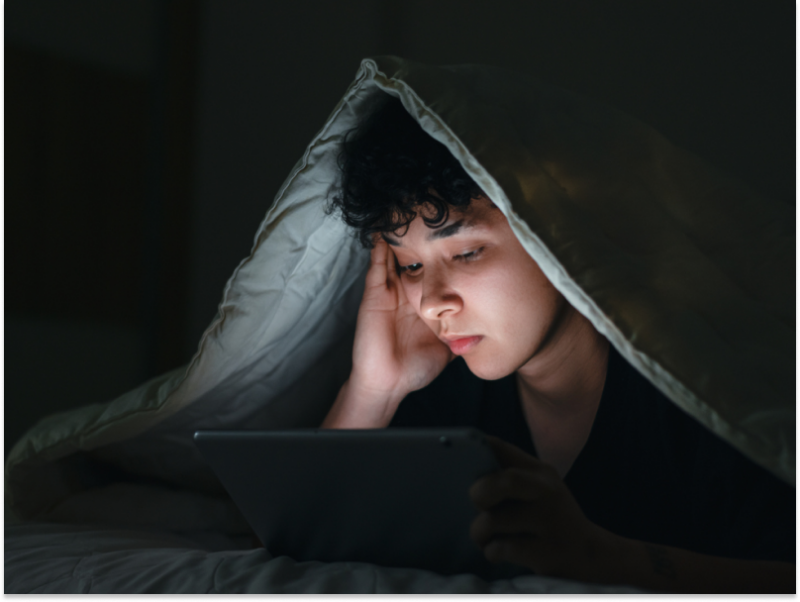 A young person looks at a screen under the covers. They are unable to sleep at night because they are anxious.