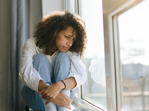 A person struggling with rejection sensitive dysphoria looks out the window while wearing jeans and a white shirt.