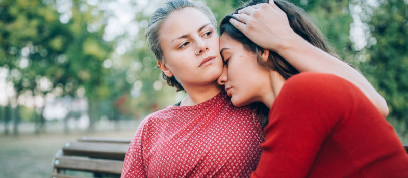 Two girls wearing red shirts embrace while sitting on a park bench. The two are in a codependent relationship.