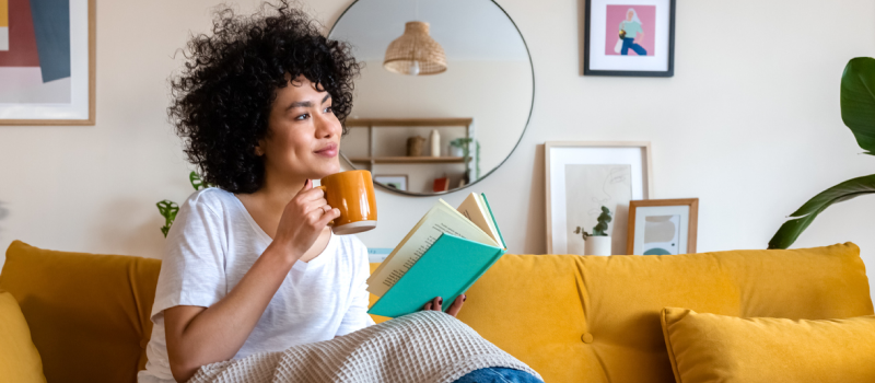 Female therapist sitting on a couch with a blanket and coffee mug. She is reading a book another therapist recommended for her.