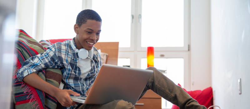 Young male in a blue and white flannel shirt with white headphones on. The teen is completing their first virtual therapy session.