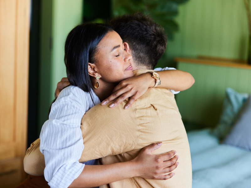 A person in a tan shirt who is struggling with grief and loss gets a hug from a person in a white shirt who is supporting him and encouraging him to attend grief counseling.