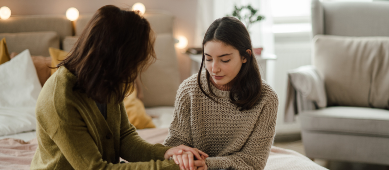 A woman in a green sweater sits on a bed next to a young person in a lighter cable knit sweater. The two hold hands and the young person looks in distress, but she is using the DBT STOP skill to manage her emotions.