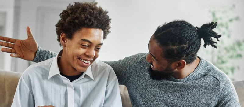 Teen with white collar sitting on a couch with father in a grey sweater. The father is proud of his son for setting mental health goals.