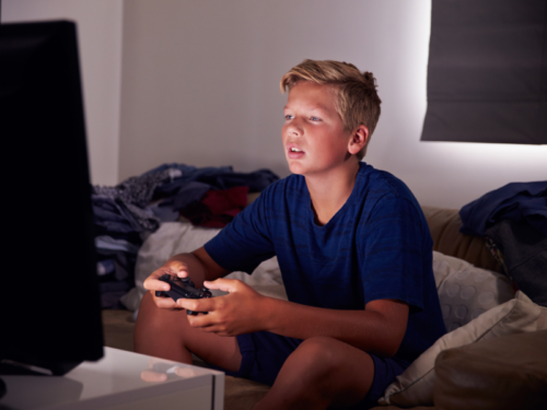 A young boy in a blue shirt plays video games late at night. He is struggling with an internet gaming disorder.