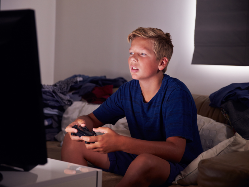 A young boy in a blue shirt plays video games late at night. He is struggling with an internet gaming disorder.