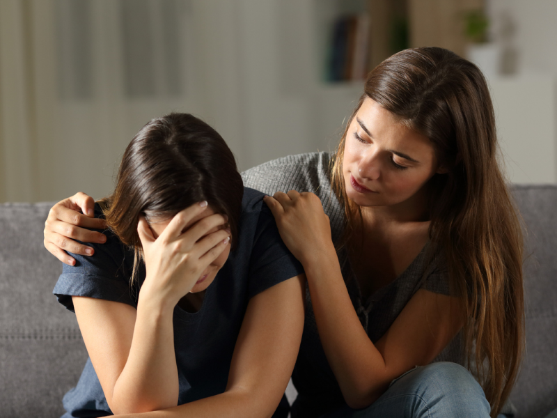 Female in grey shirt on couch with sibling in black shirt. The girl is comforting her sibling who is having anxiety.