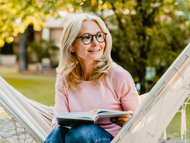 A young woman who is a mother to a teenager sitting on a hammock in a pink shirt and blue jeans. She is reading a parenting book.