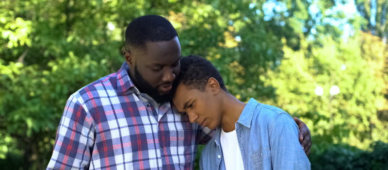 A young male in a blue shirt is being comforted by his father in a plaid shirt. The teenager is suffering from anxiety and experiencing migraines.