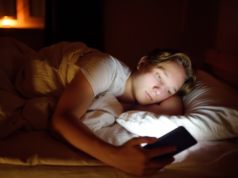 Young male laying in bed with a white shirt on their phone. The young male is