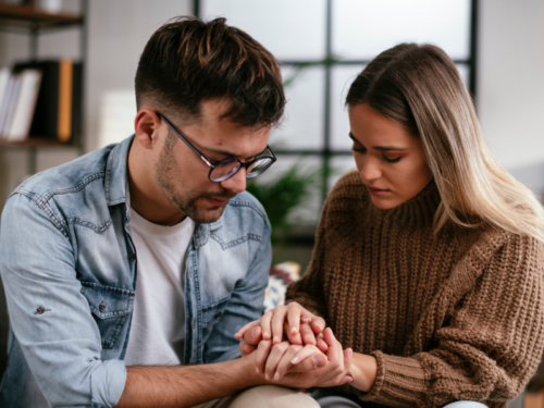 A young couple sits, comforting each other on the couch. The young woman is experiencing postpartum depression after a miscarriage.