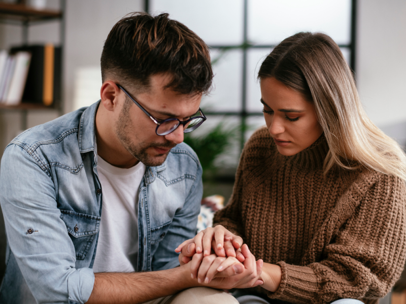 A young couple sits, comforting each other on the couch. The young woman is experiencing postpartum depression after a miscarriage.