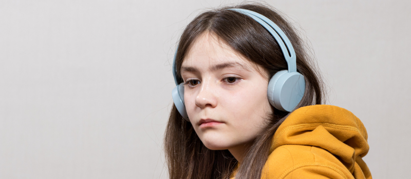 A female teenager in a yellow sweatshirt and blue headphones. She suffers from migraines and depression.