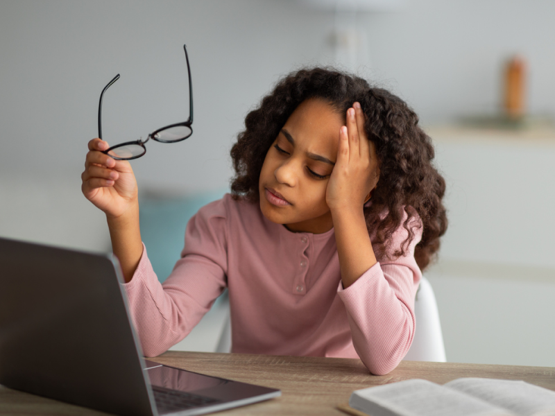 A female teenager in a pink shirt sitting at her desk. She is experiencing a migraine and wonders if it is related to her anxiety disorder.