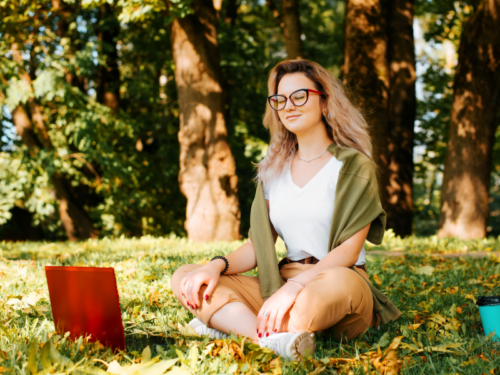 A female young adult in a green sweater and white shirt sitting outside on the grass. She is using somatic exercises to improve her mental health.