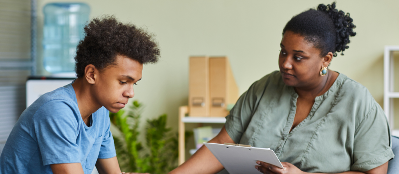 Male teenager in a blue shirt at weekly therapy with a female therapist in a green shirt. He is stepping up in care from weekly therapy to Intensive Outpatient Programming (IOP)>