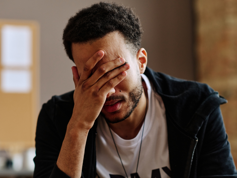 A young male veteran in a white shirt and a black jacket. The male has his head in his hand and is suffering from war trauma.