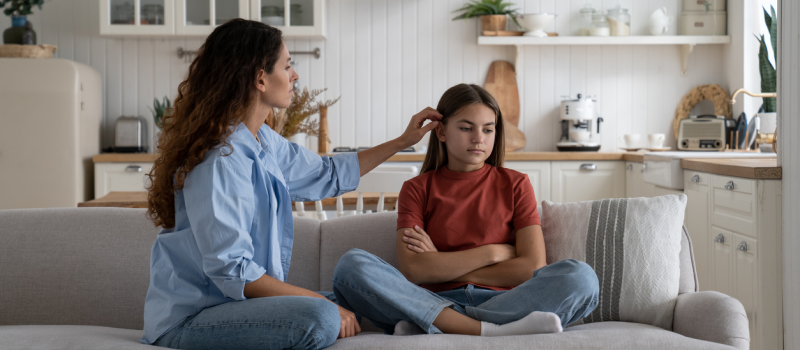 A woman in a blue shirt trying to comfort her daughter in a red shirt. They sit together on a couch. The mother eggshell parents due to her unpredictable behavior and outbursts.