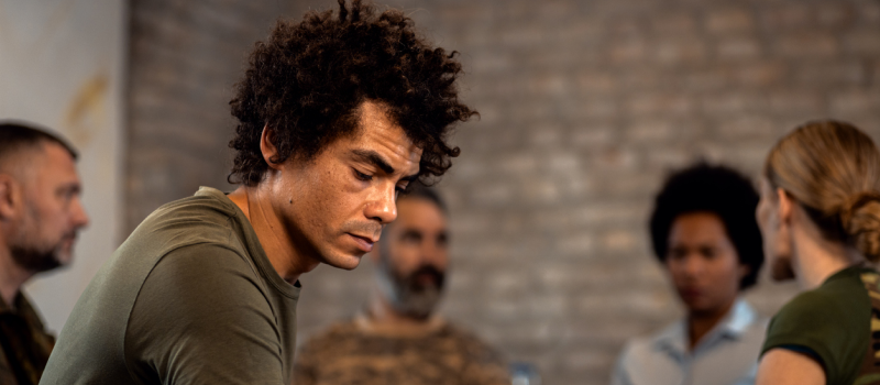 A male veteran in a green shirt sits with other veterans in a room. The male has war trauma as a psychological consequence of war.