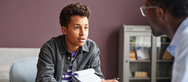 Male teenager in a purple shirt with a grey jacket talking to an older male therapist. The teen is being treated for quiet borderline personality disorder.
