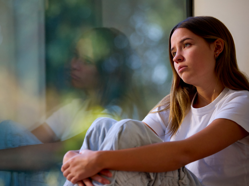 Young female in a white shirt and sweatpants looking outside of her window. She is being treated for depression.