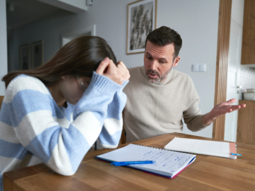 A father in a tan shirt sits with his daughter in a white and blue sweater at a table. He is having an unpredictable outburst and is eggshell parenting.