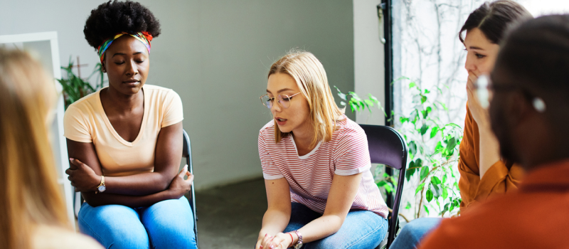 A young woman in a striped shirt sits in a circle of other young individuals. She is in a support group for pregnancy loss due to experiencing Postpartum Depression after a miscarriage.