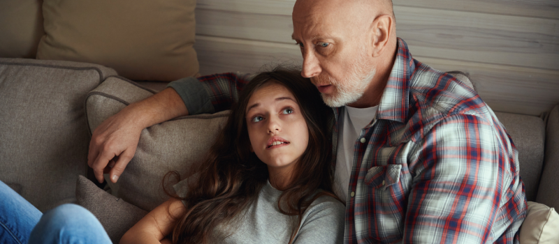 Young female in grey shirt discussing with her father in plaid shirt that her depression is making her sick.