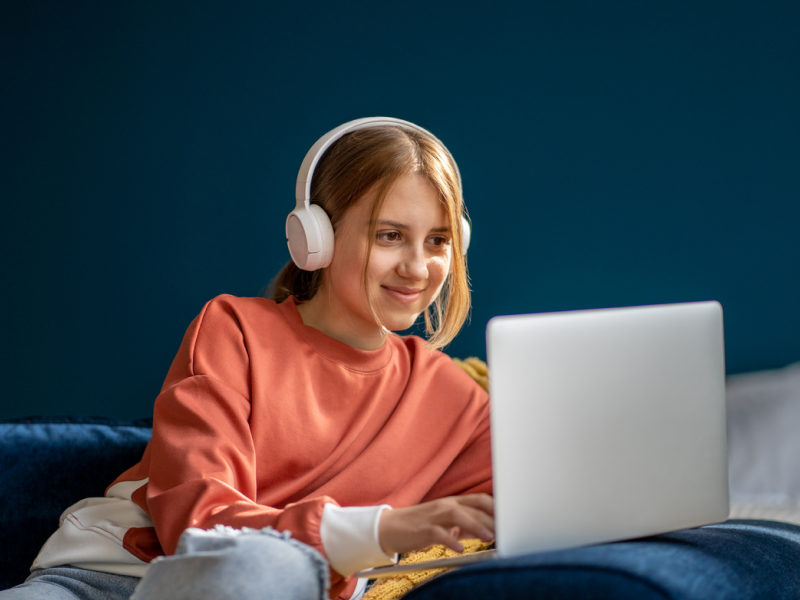 A female teenager sits in her room in an orange shirt. She is in virtual therapy, where the program uses measurement-based care.