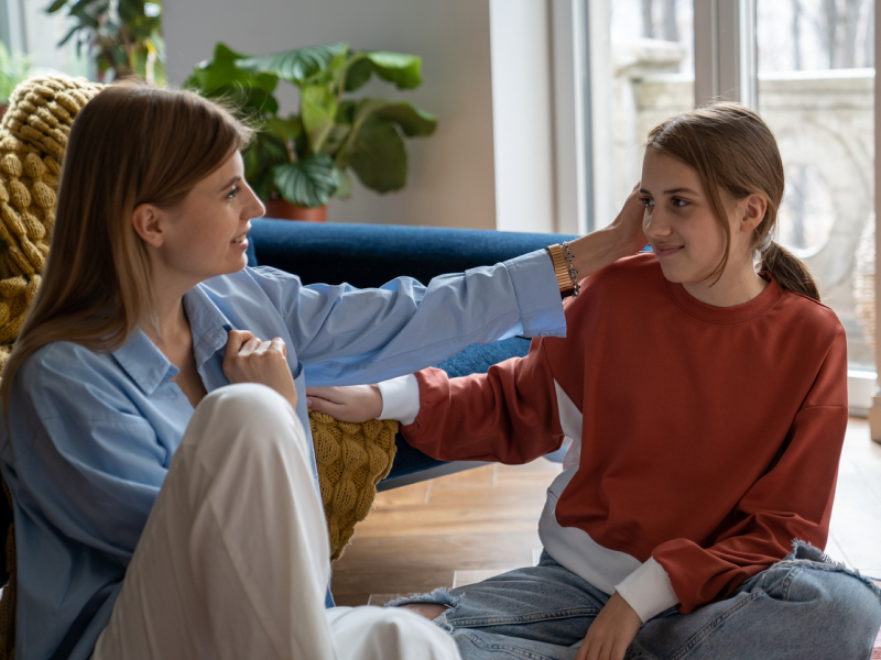 A mother and daughter sitting on the ground in their living room. The mother is supporting the daughter who is having signs of paranoid personality disorder.
