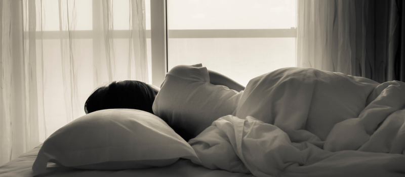 Young person laying in their bed looking towards the window in a white shirt and white bed set. The person is "bed rotting", spending excessive time in bed.