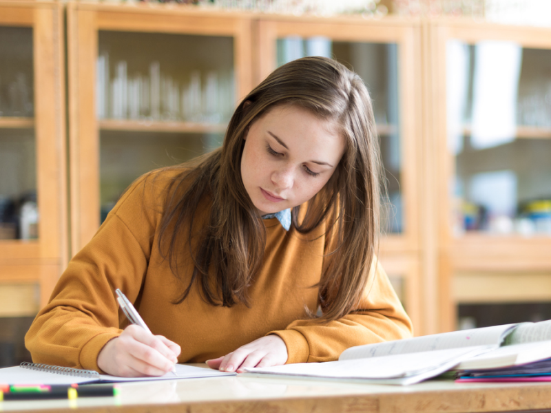 A female high schooler sitting at her desk doing homework in an orange sweatshirt. She has ADHD and has been learning the best tips for her to focus with ADHD.