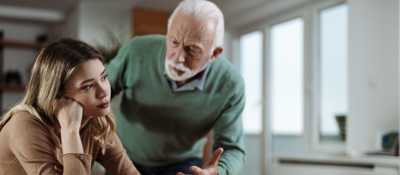 A woman wearing a brown shirt looks into the distance as her father, who stands in the background in a green shirt and has narcissistic personality disorder, tries to use manipulation tactics to get her to do something he wants.