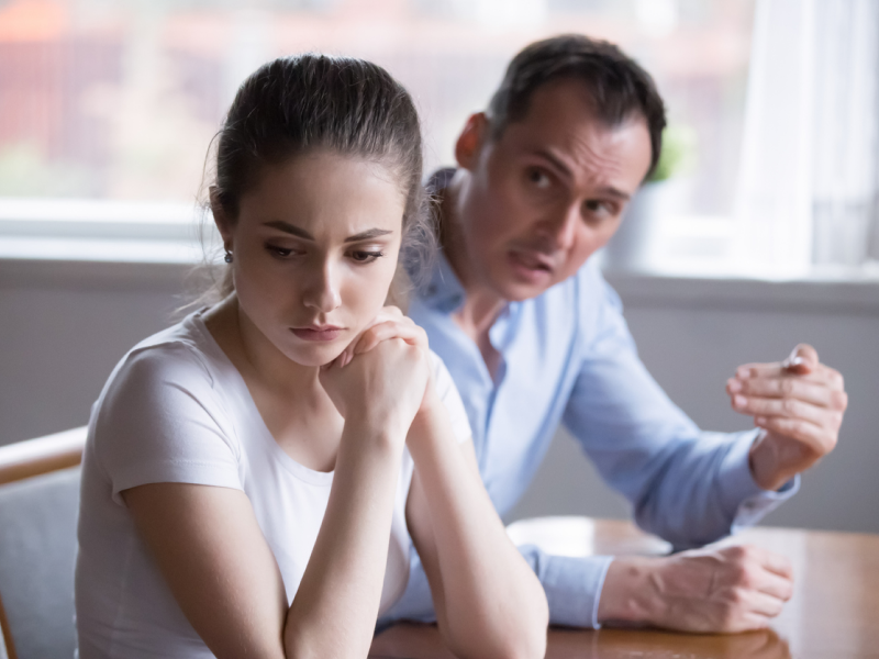 A father, who has narcissistic personality disorder, berates his daughter who sits quietly grasping her hands and looking down at a table.