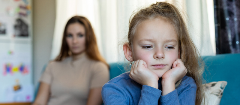 A young girl rest her face on her hand as her narcissistic mother sits in the background looking at her.
