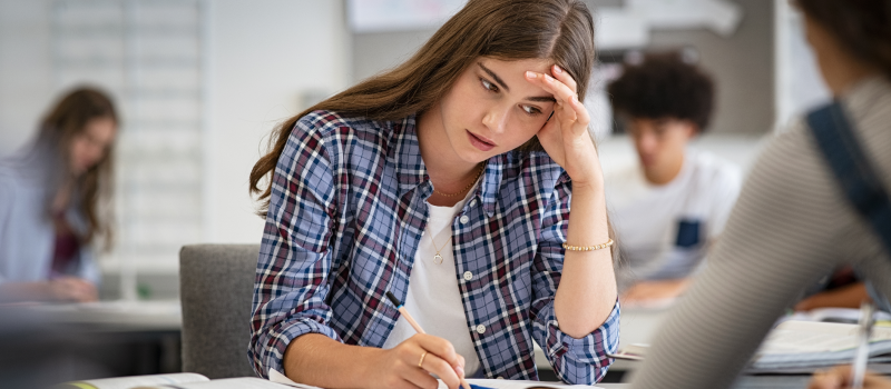 A student in a plaid button down holds her forehead as she works on an assignment. She is able to focus at school since she has taken care of her physical and emotional safety by eating enough food and practicing self-care.