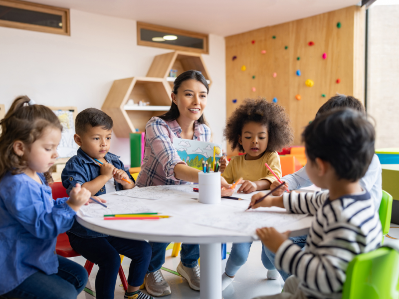 A teacher sits at a table with students as they return to school. The teacher is instructing a back-to-school art activity to reduce back-to-school mental health struggles.