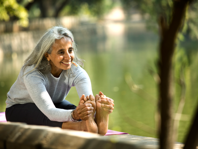A person with grey hair wearing yoga clothes stretches next to a pond after completing a mindfulness meditation exercise.